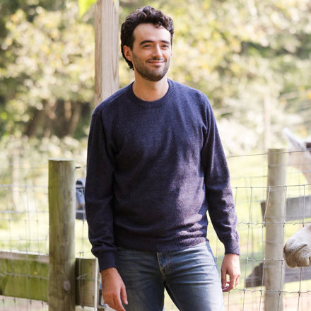 Men's Classic Crewneck 100% Baby Alpaca Sweater. A man wearing a blue baby alpaca crewneck sweater stands in front of a farm fence.