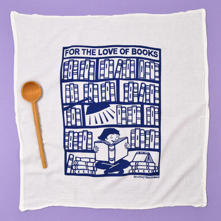 White towel with blue graphic of a person reading a book between bookshelves, accompanied by a wooden spoon on a purple background.