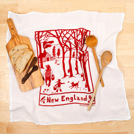 Tea towel with red New England design on a white towel, surrounded by bread, a cutting board, and wooden spoons.